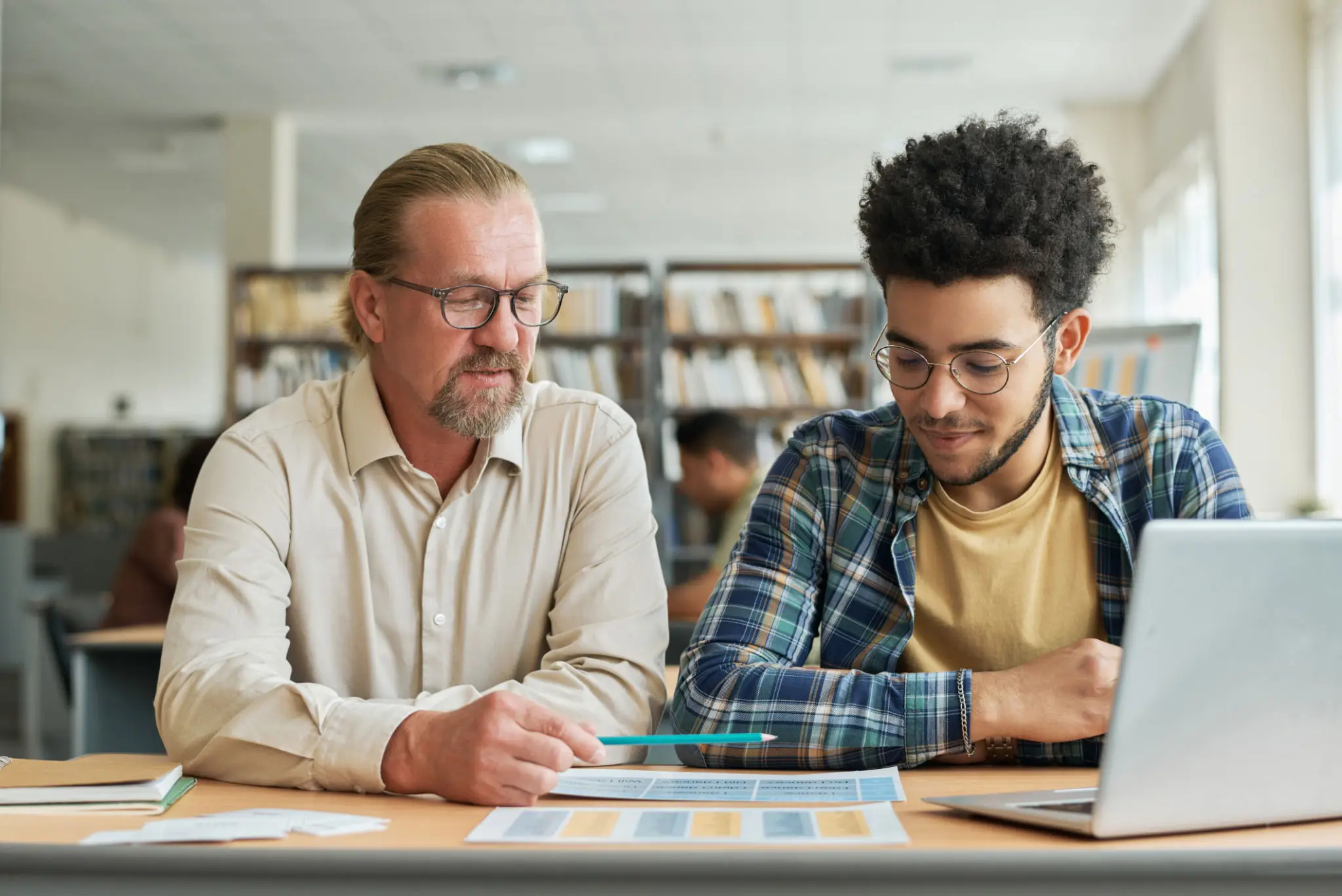 A teacher guides a student in a library, reviewing a book together.